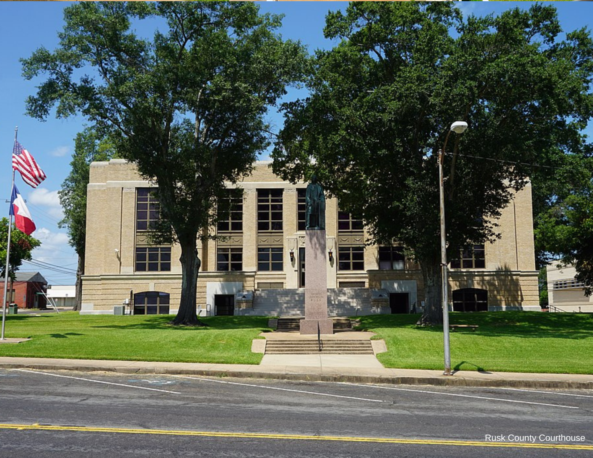 Stone courthouse with flags and a war memorial in front, seen across a street under trees.