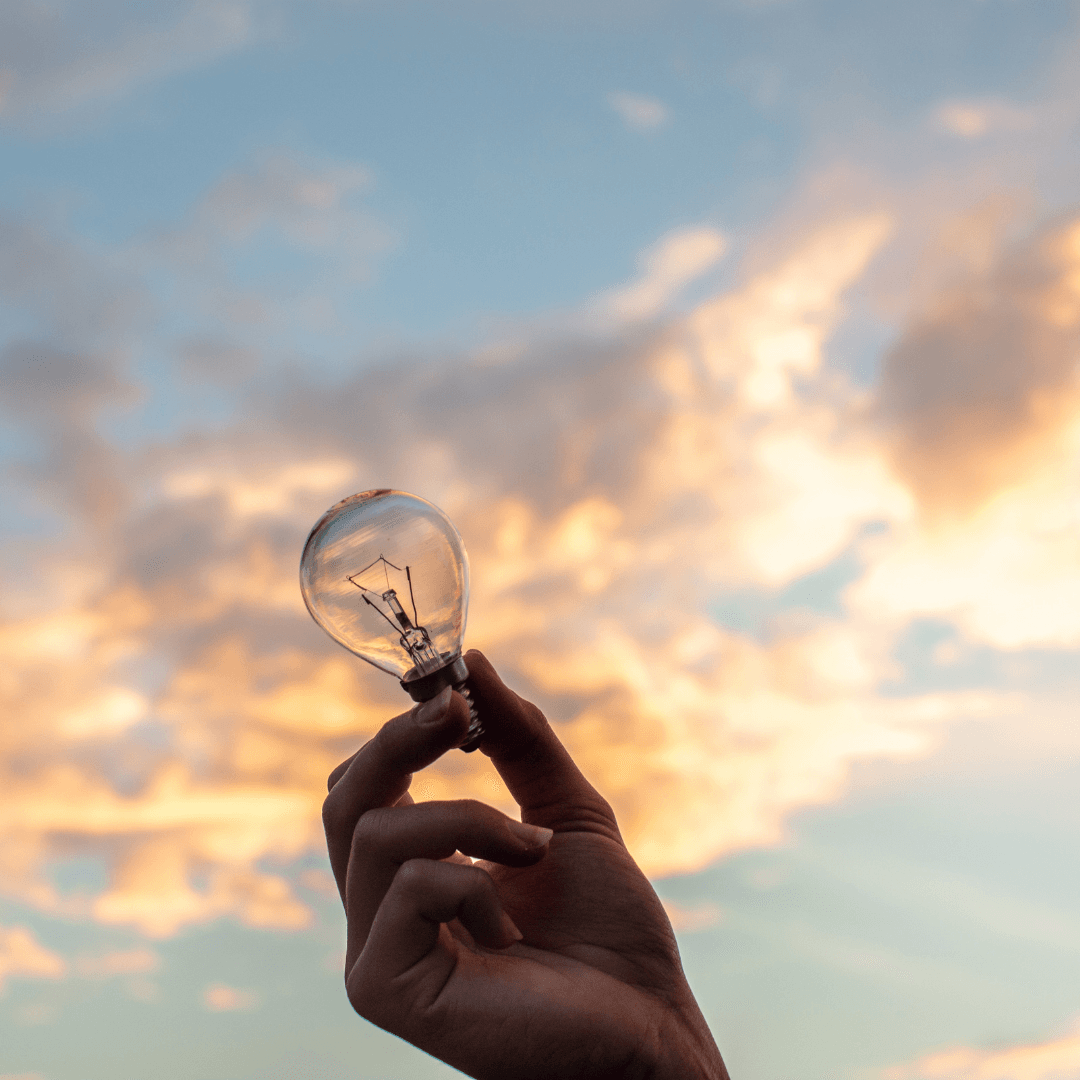 Hand holding a light bulb against a pastel sunset sky