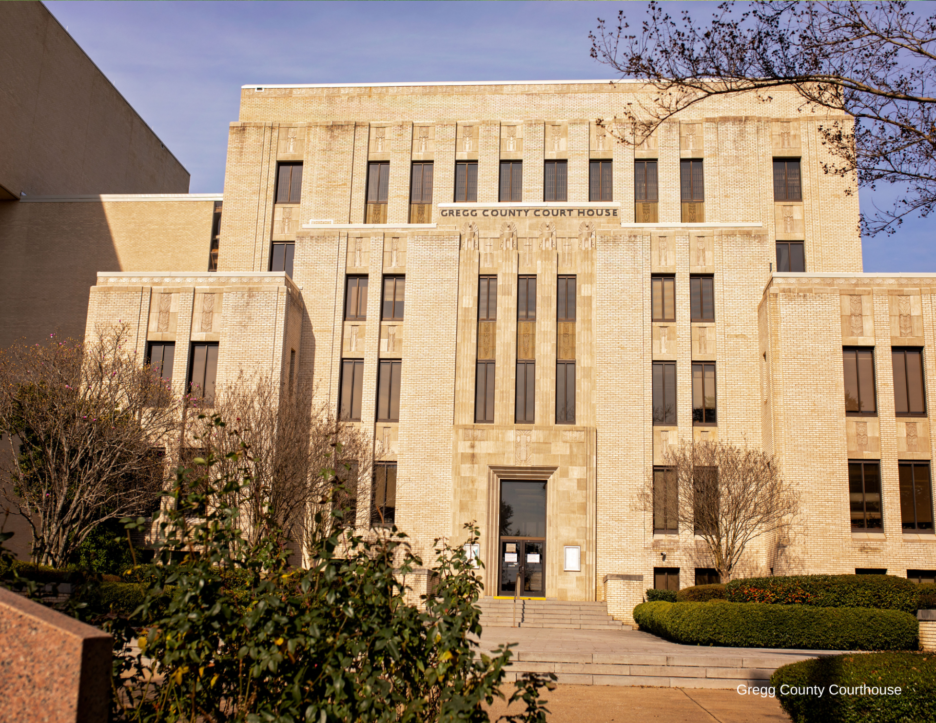 Beige stone campus building with central entrance, windows, and trees in front under a clear sky
