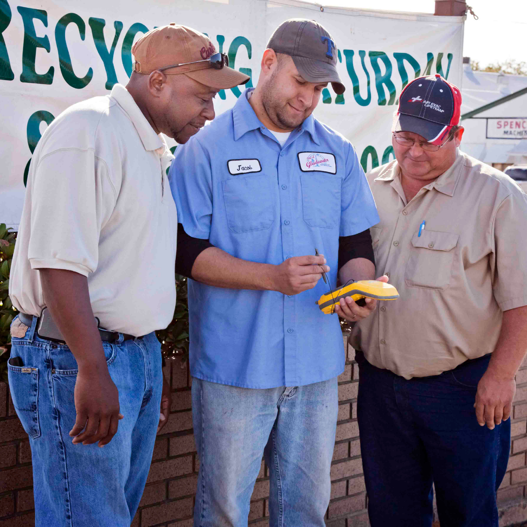 Three men in work clothes review a yellow item together at an outdoor recycling center.