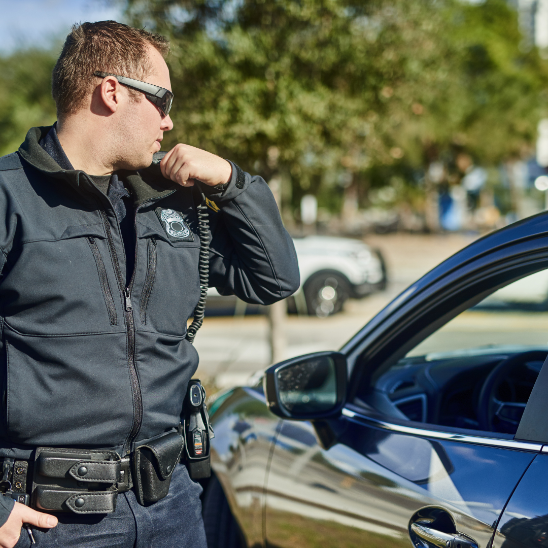 Police officer standing by a blue car, talking on a radio in a sunny parking lot