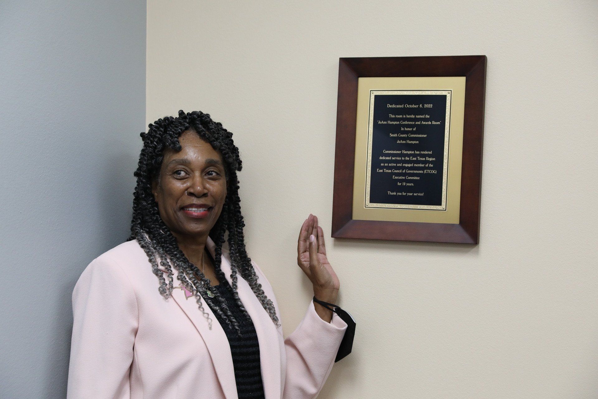 A woman in a pink jacket is standing next to a framed plaque on a wall.