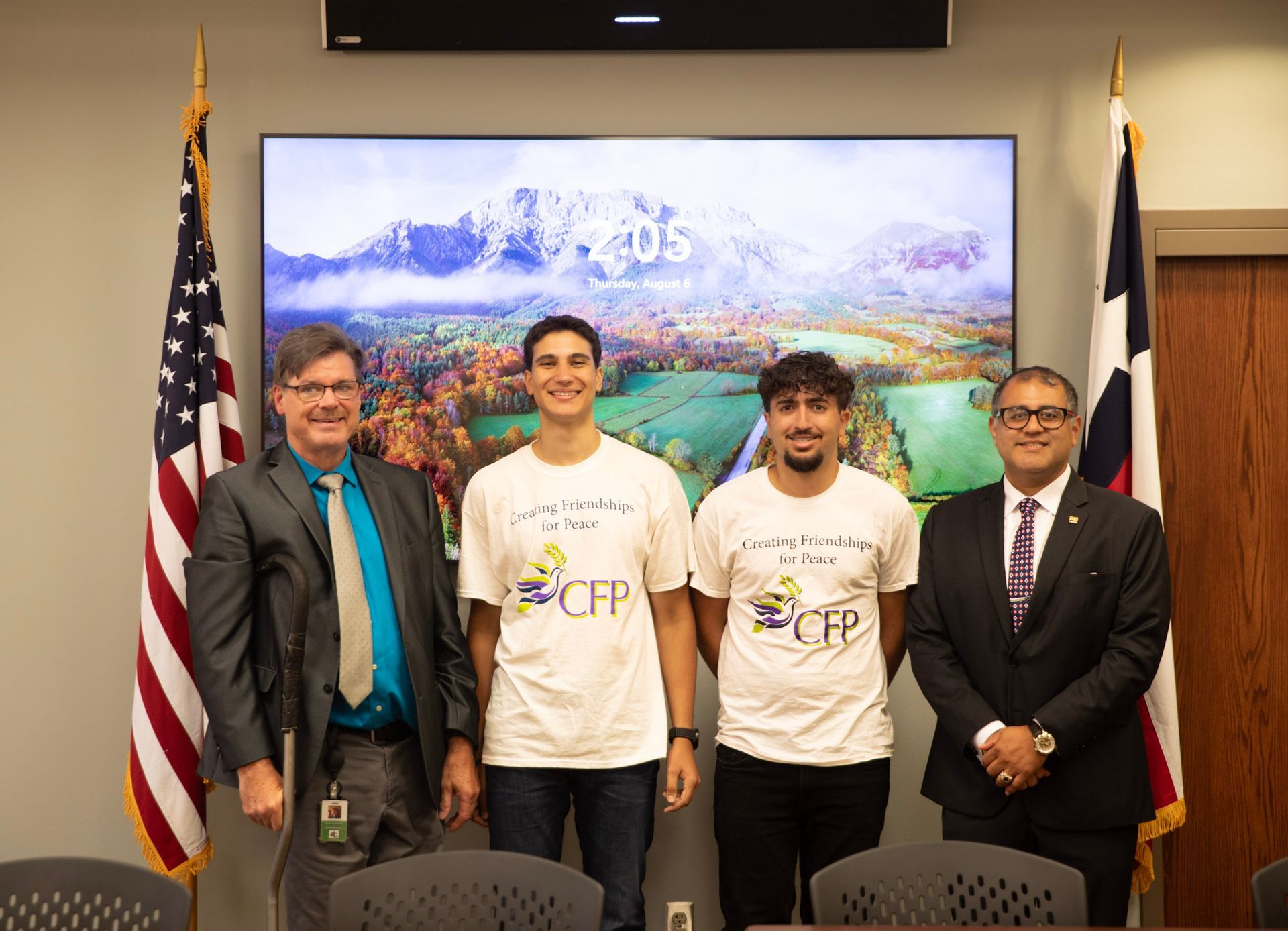 Four people posing indoors in front of a landscape screen, with U.S. and Texas flags on either side.