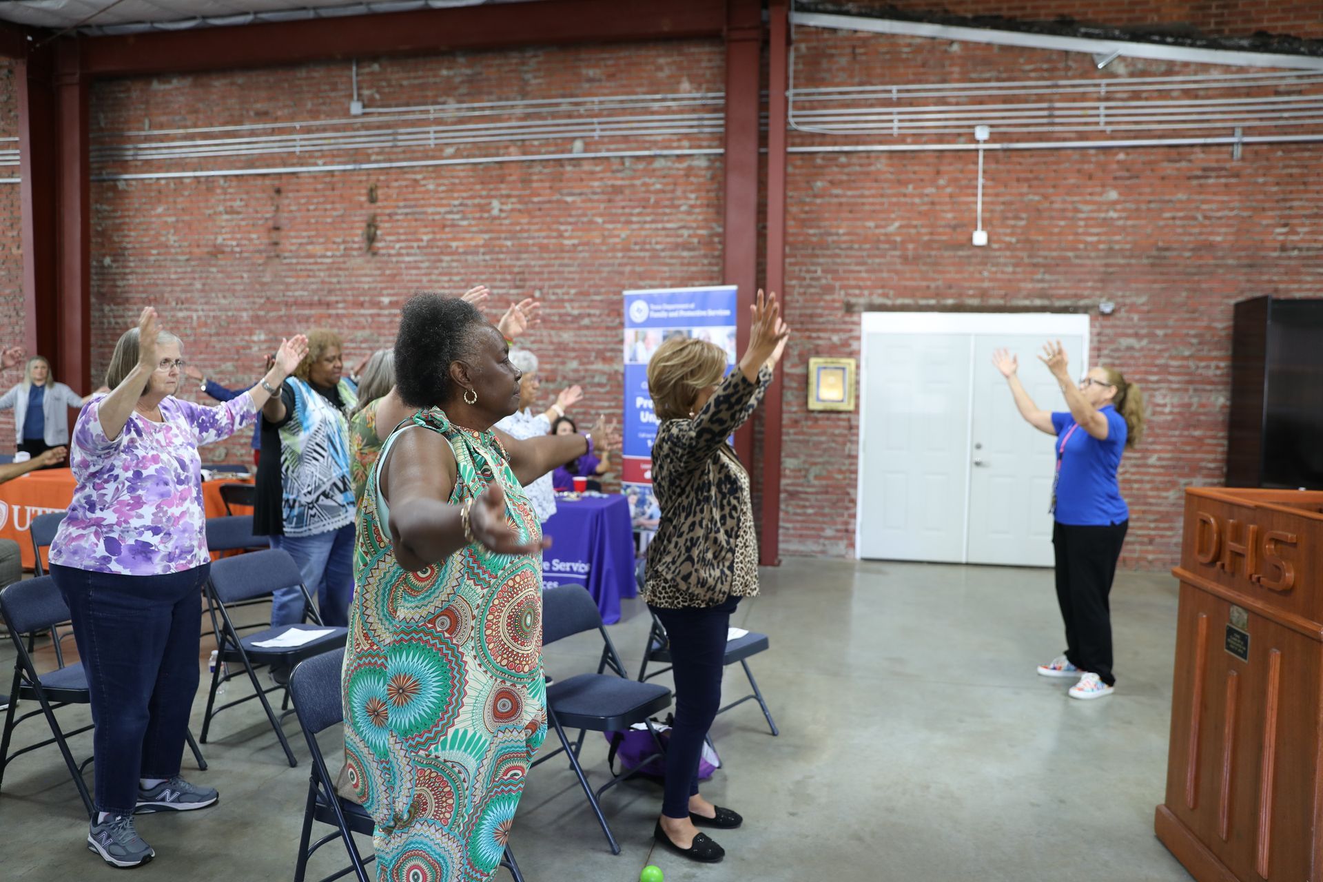 Group of people stretching arms up in a room. A woman leads the stretch, standing by a lectern.