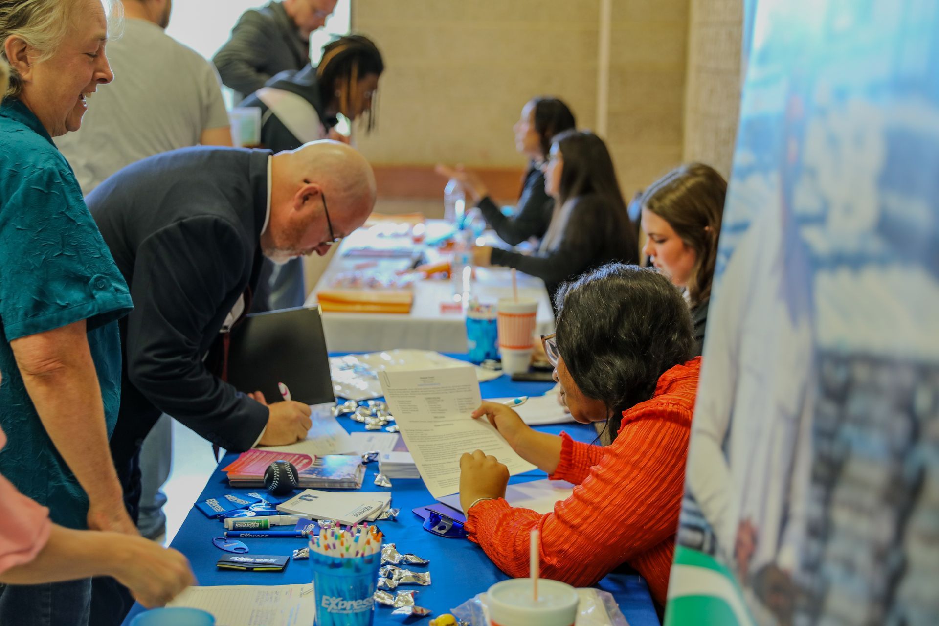 People signing documents at a table; others nearby. Pens, paperwork, and blue table cloth.