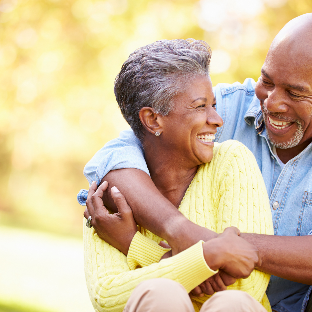 Smiling couple hugging outdoors, wearing yellow and blue sweaters against a bright blurred background