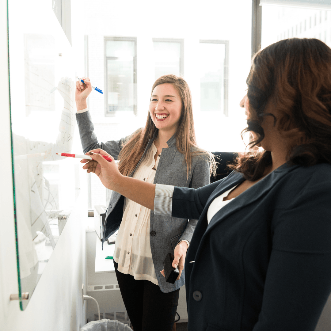 Two coworkers point at notes on a glass whiteboard in a bright office.
