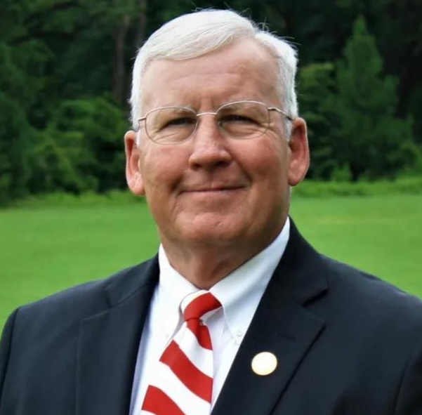 Man in a dark suit with red striped tie, standing outdoors on a green lawn with trees behind him.