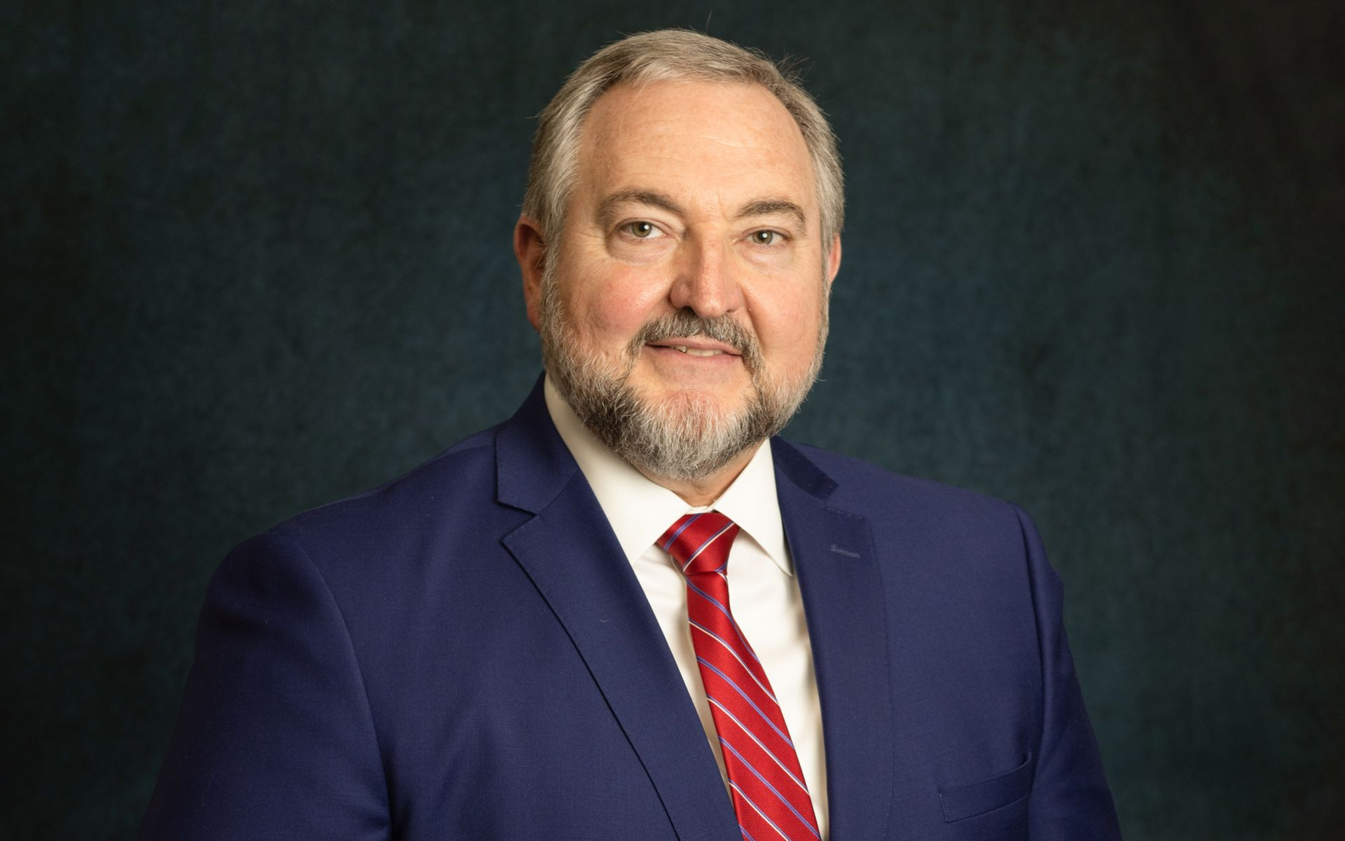 Portrait of a man in a navy suit and red striped tie against a dark background