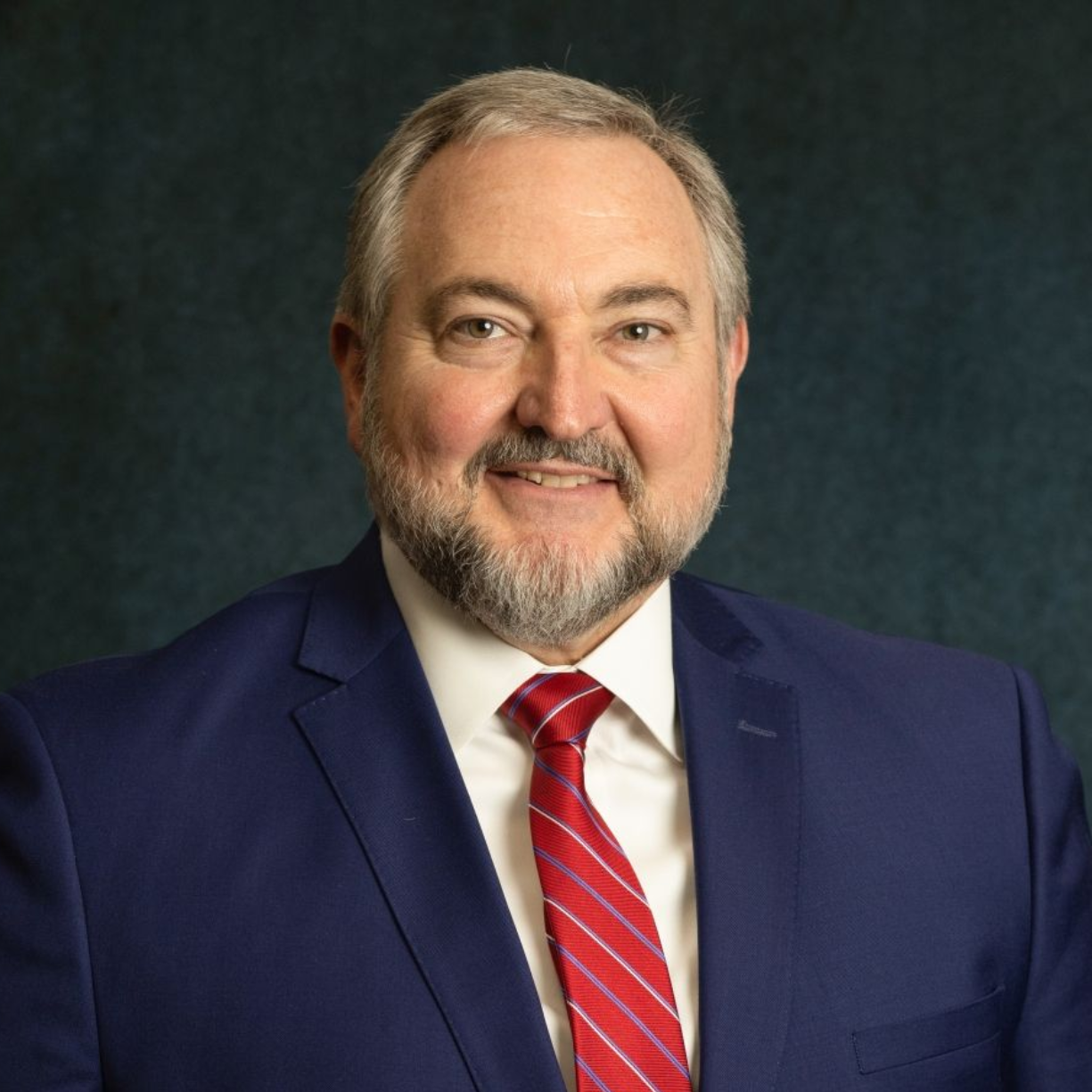 Studio headshot of a smiling man in a navy suit and red striped tie, against a dark background
