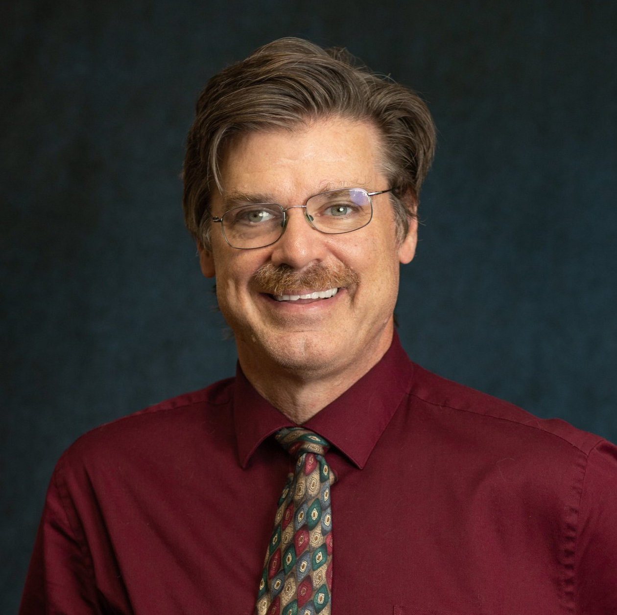 Portrait of a smiling man in glasses, red shirt, and patterned tie against a dark studio background
