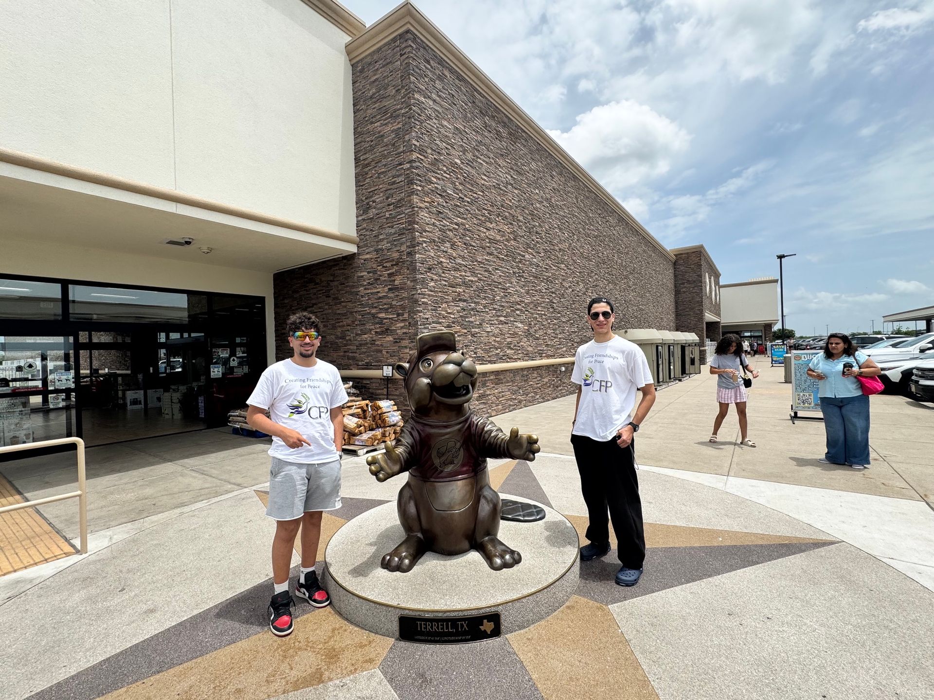 Two people posing with a bronze bear statue outside a store on a sunny sidewalk.