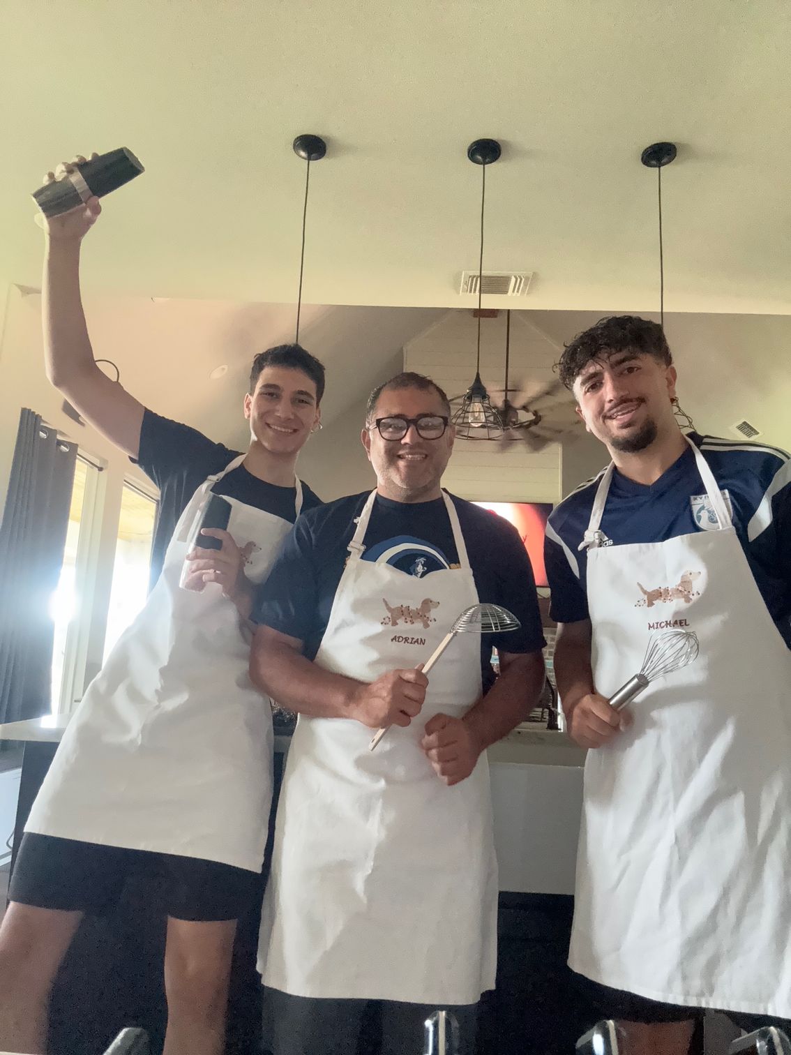 Three people in aprons smiling and posing in a kitchen, one holding up a bottle.