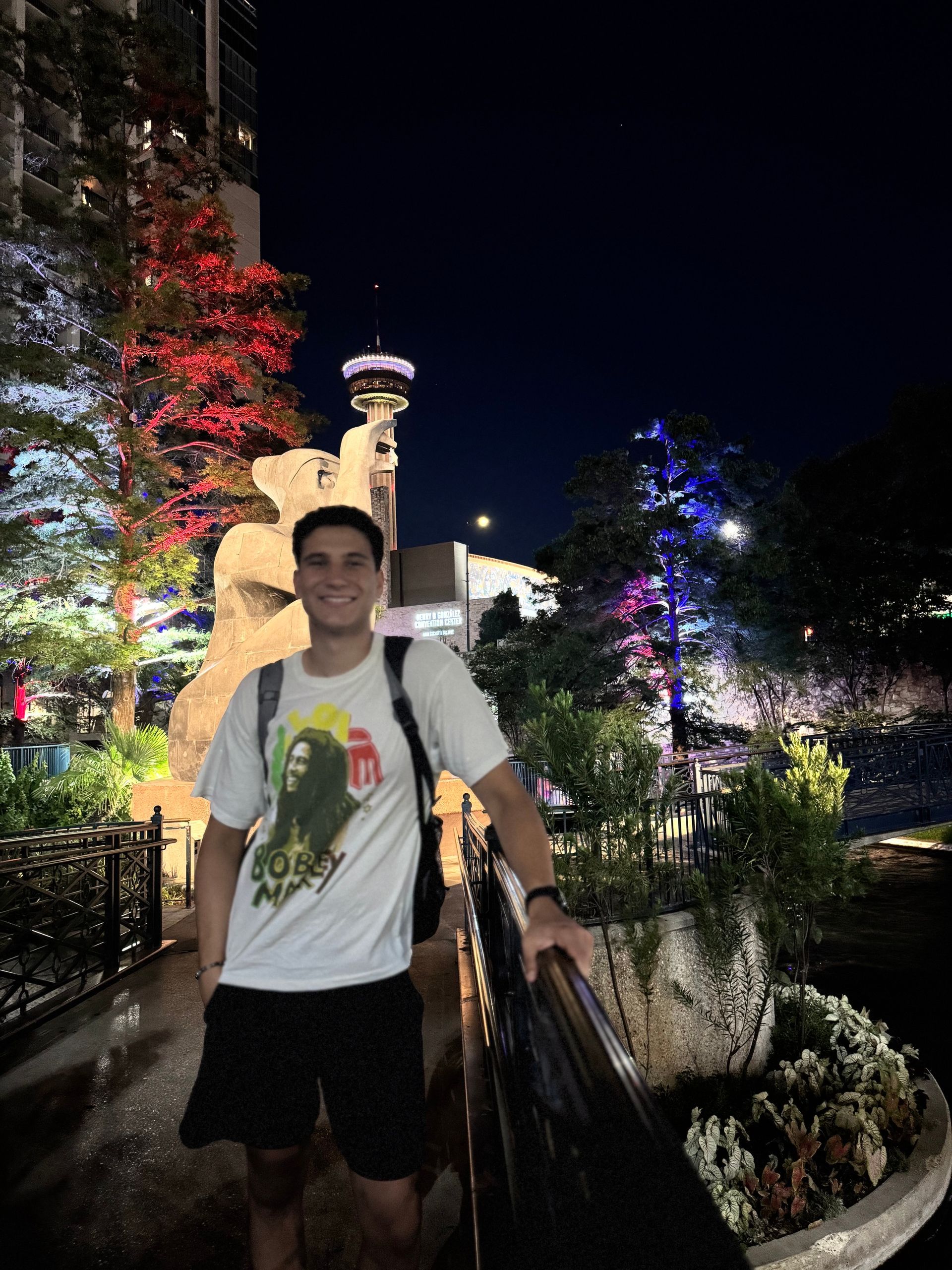 Person smiling with a backpack at a brightly lit night garden near a lit tower