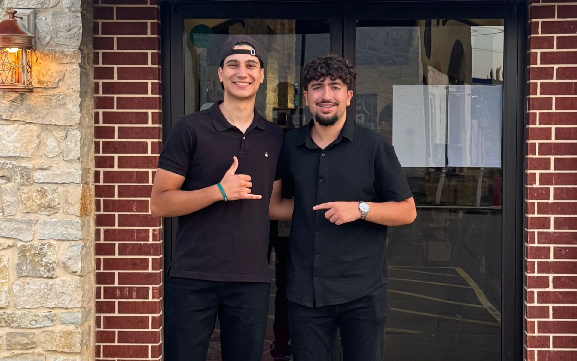 Two people in black shirts smiling outside a brick storefront, one giving a thumbs-up.