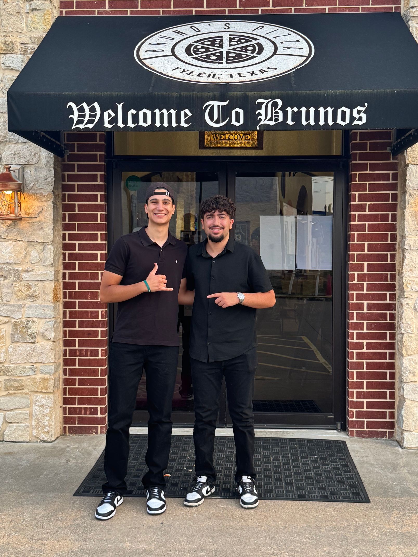 Two people standing outside Bruno’s restaurant entrance, wearing black outfits and smiling.