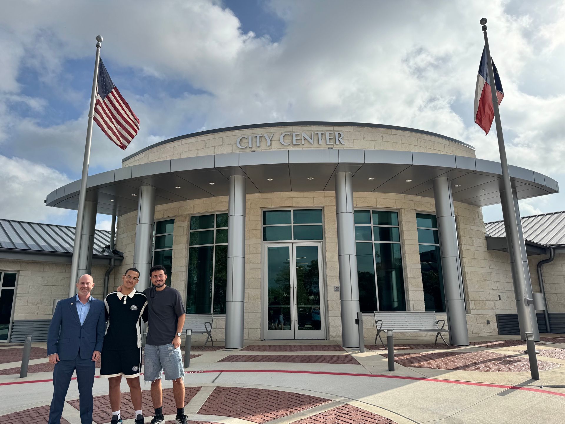 Three men standing outside a circular civic building with U.S. and Texas flags flying above the entrance.