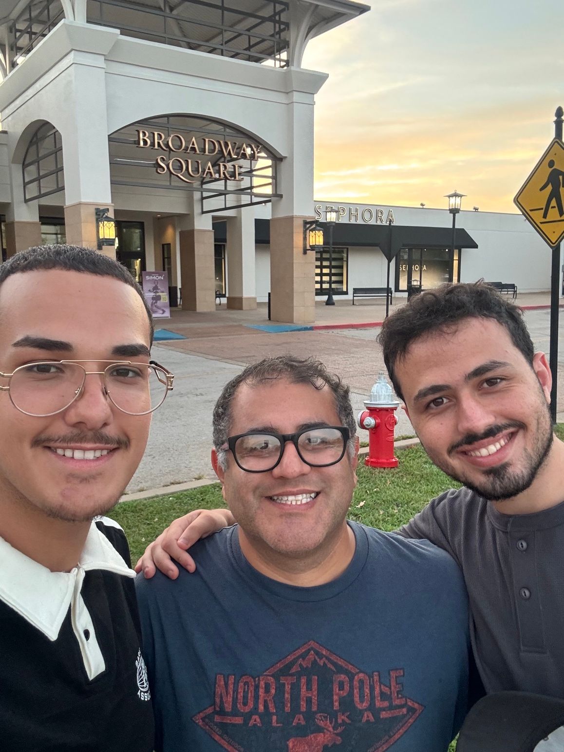 Three smiling people posing outside a building at sunset with a visible “North Pole” sign.