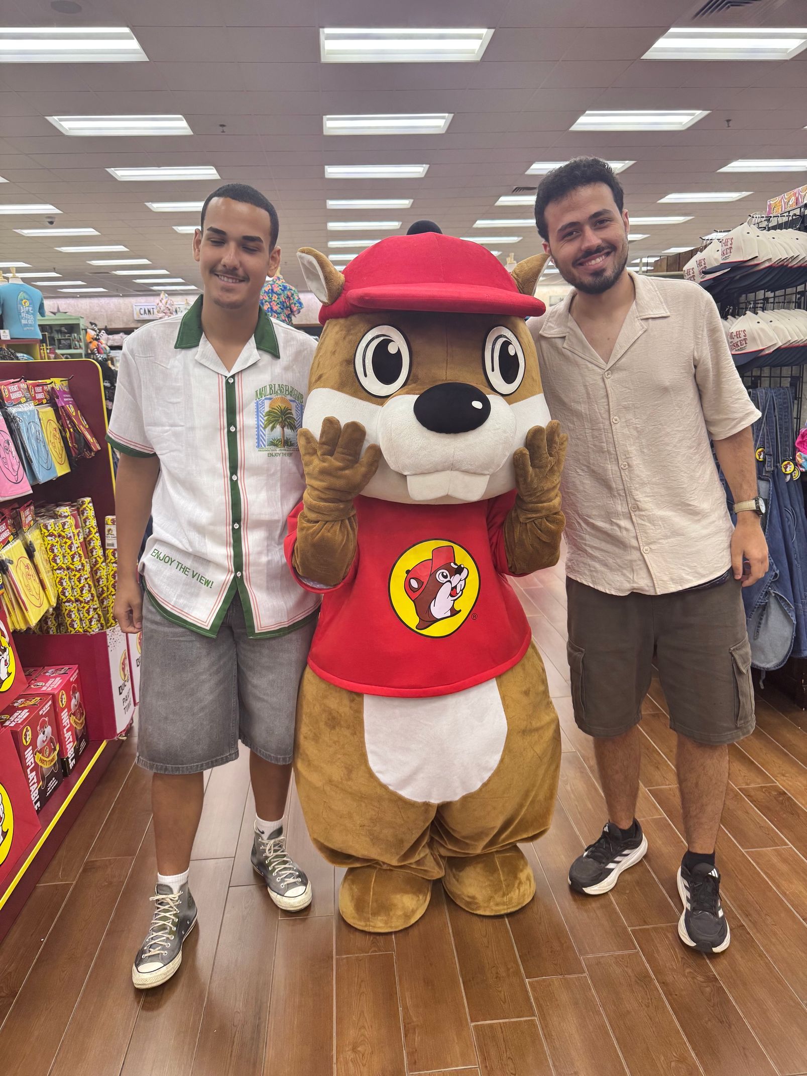 Two men posing with a red-hatted mascot in a store aisle, all smiling.