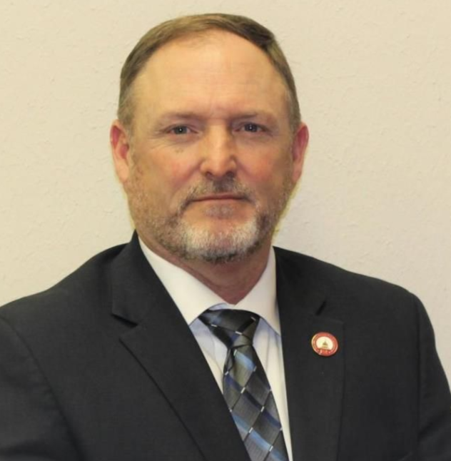 Man in a dark suit and striped tie with a lapel pin, posing against a plain beige background.