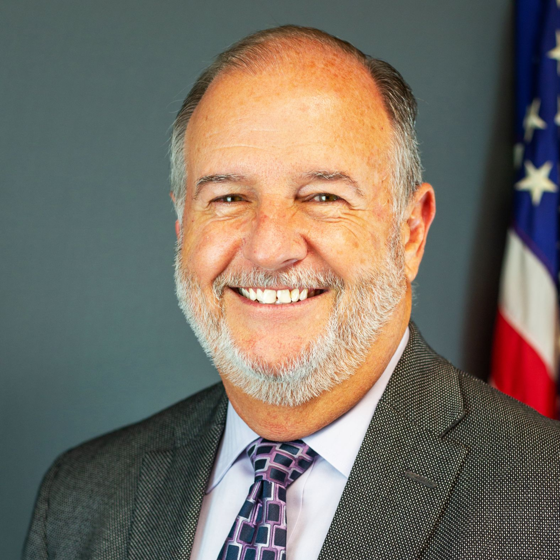 Portrait of a smiling man in a suit and tie, with an American flag in the background