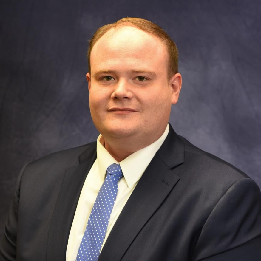 Portrait of a man in a dark suit and blue tie against a mottled blue studio backdrop