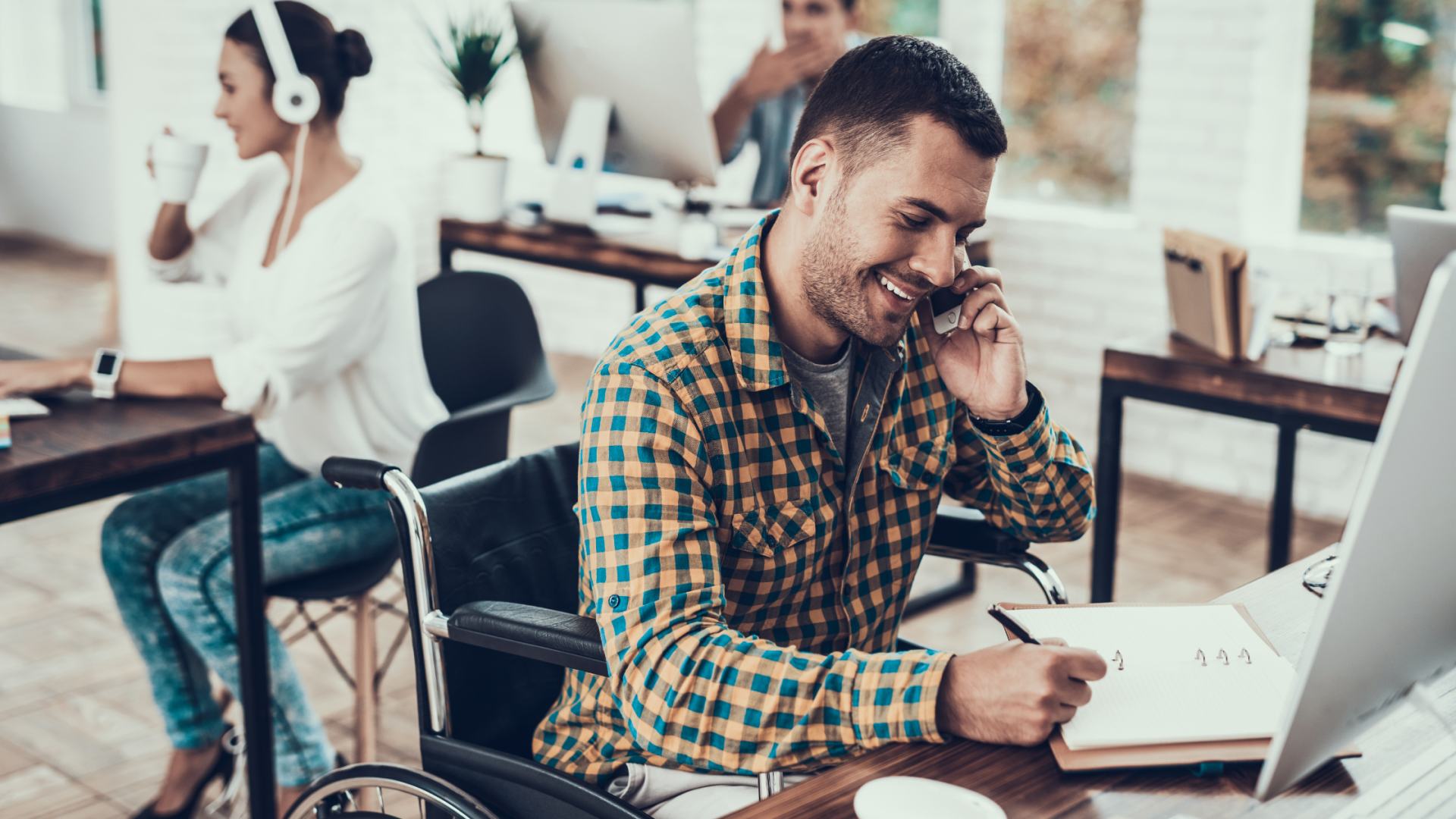 A man in a wheelchair is sitting at a desk talking on a cell phone.