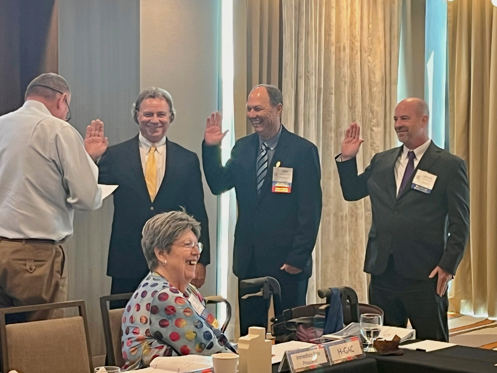 Group of men taking oath with hands raised, led by a person holding a paper, while a woman sits smiling. Conference room.