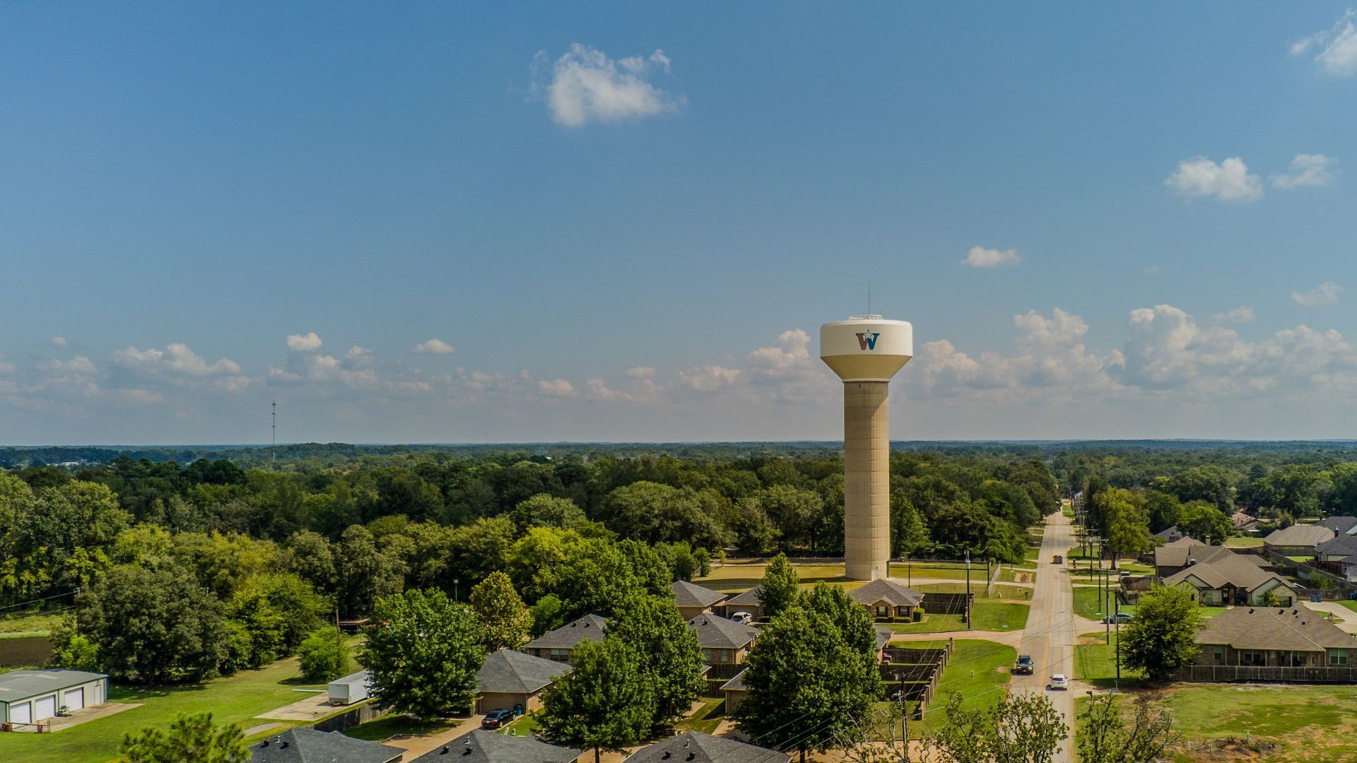 A water tower stands prominently in a rural residential neighborhood, surrounded by lush green trees under a blue sky.