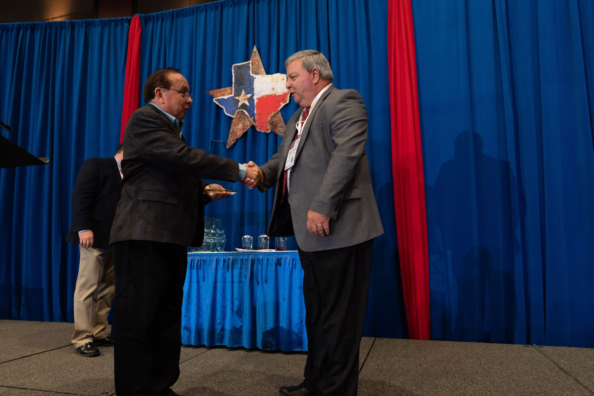 Two men shaking hands in front of a blue curtain with a texas star on it
