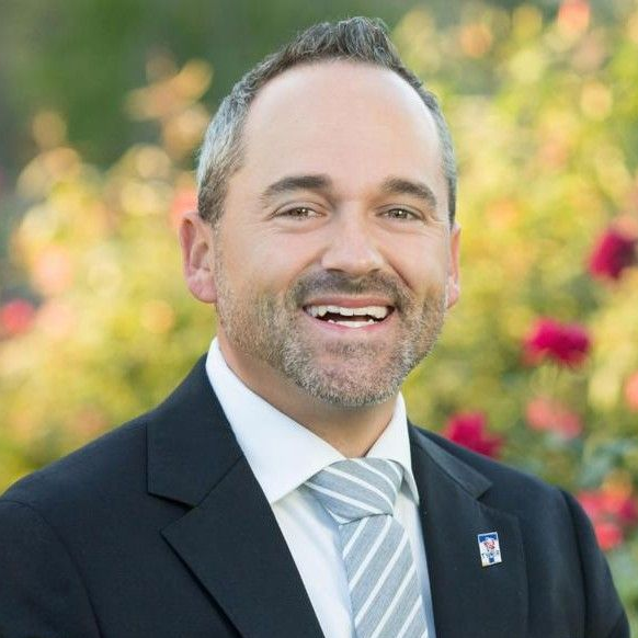 Smiling man in a gray suit and blue tie standing outdoors in front of a building