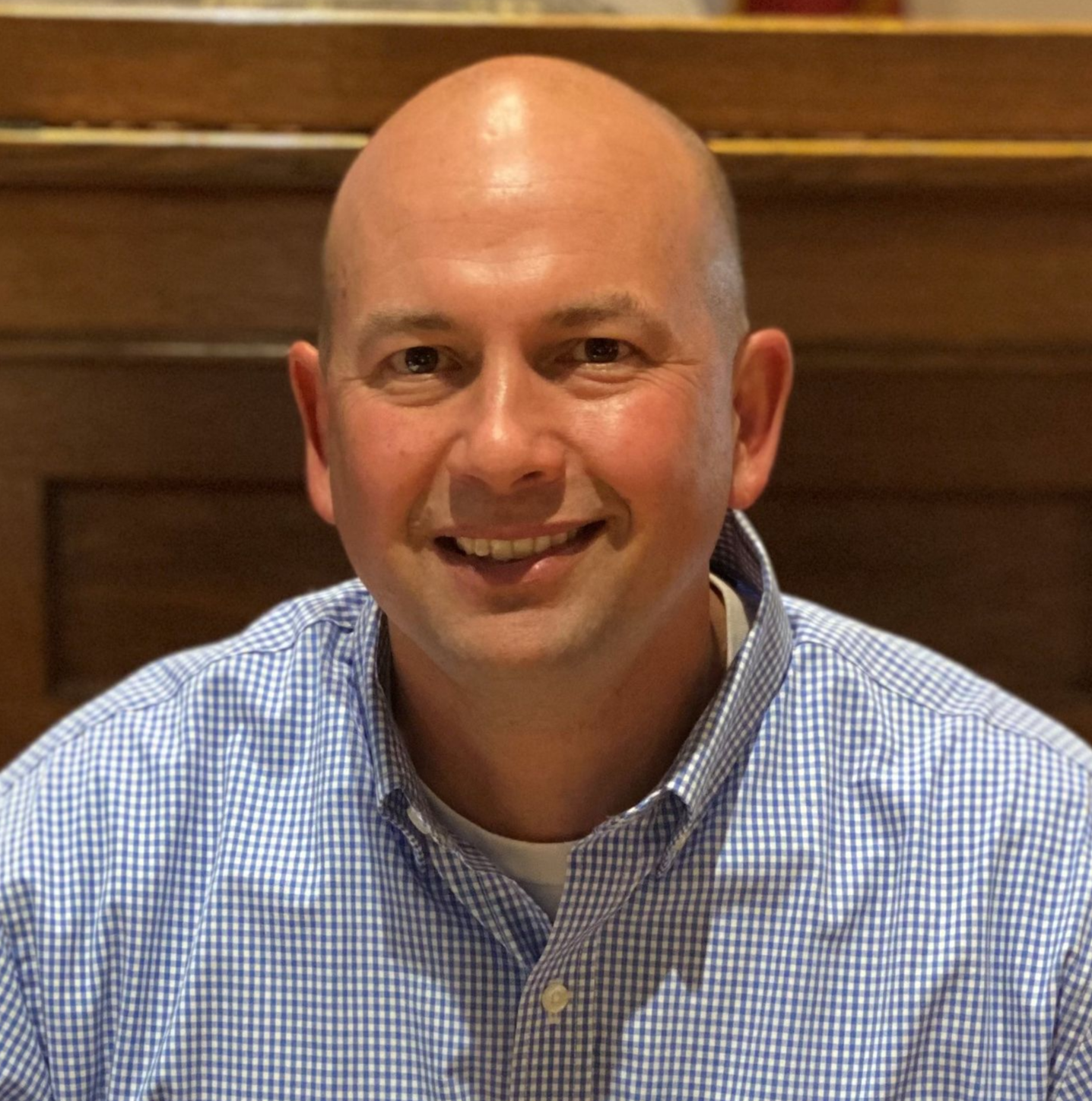 Smiling bald man in a blue checkered shirt seated indoors against a wooden backdrop