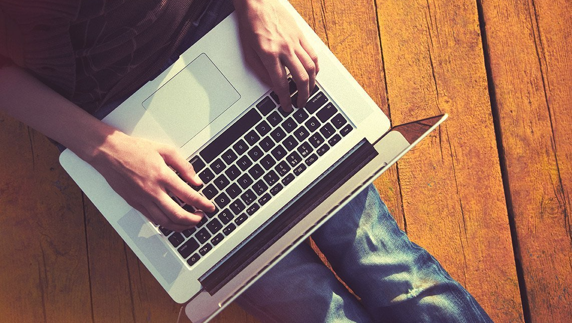 A person is typing on a laptop computer while sitting on a wooden floor.