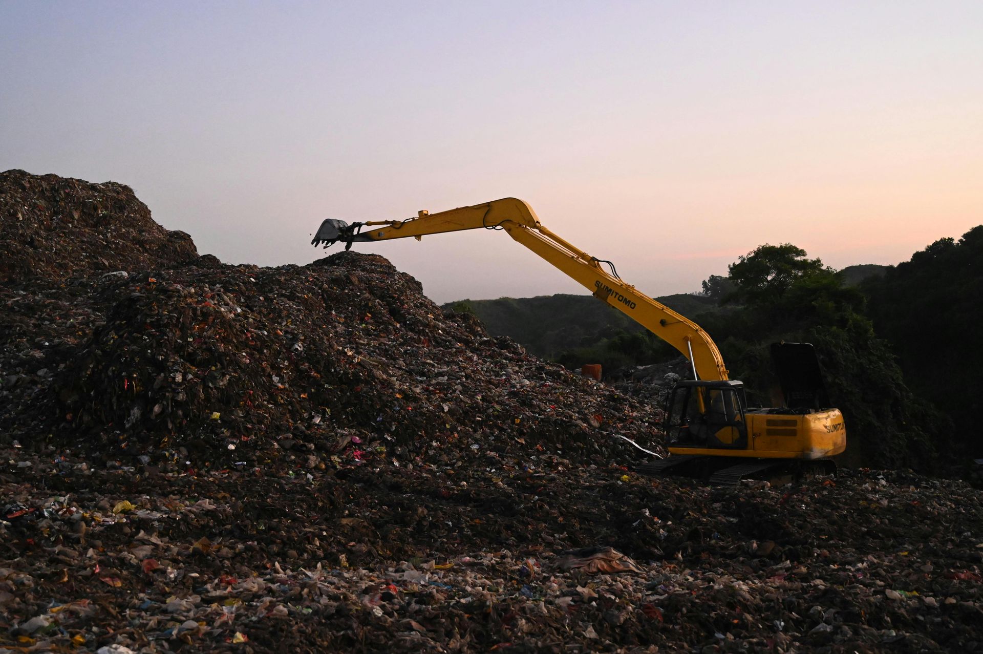 Yellow excavator on a mountain of trash, against a dusk sky.