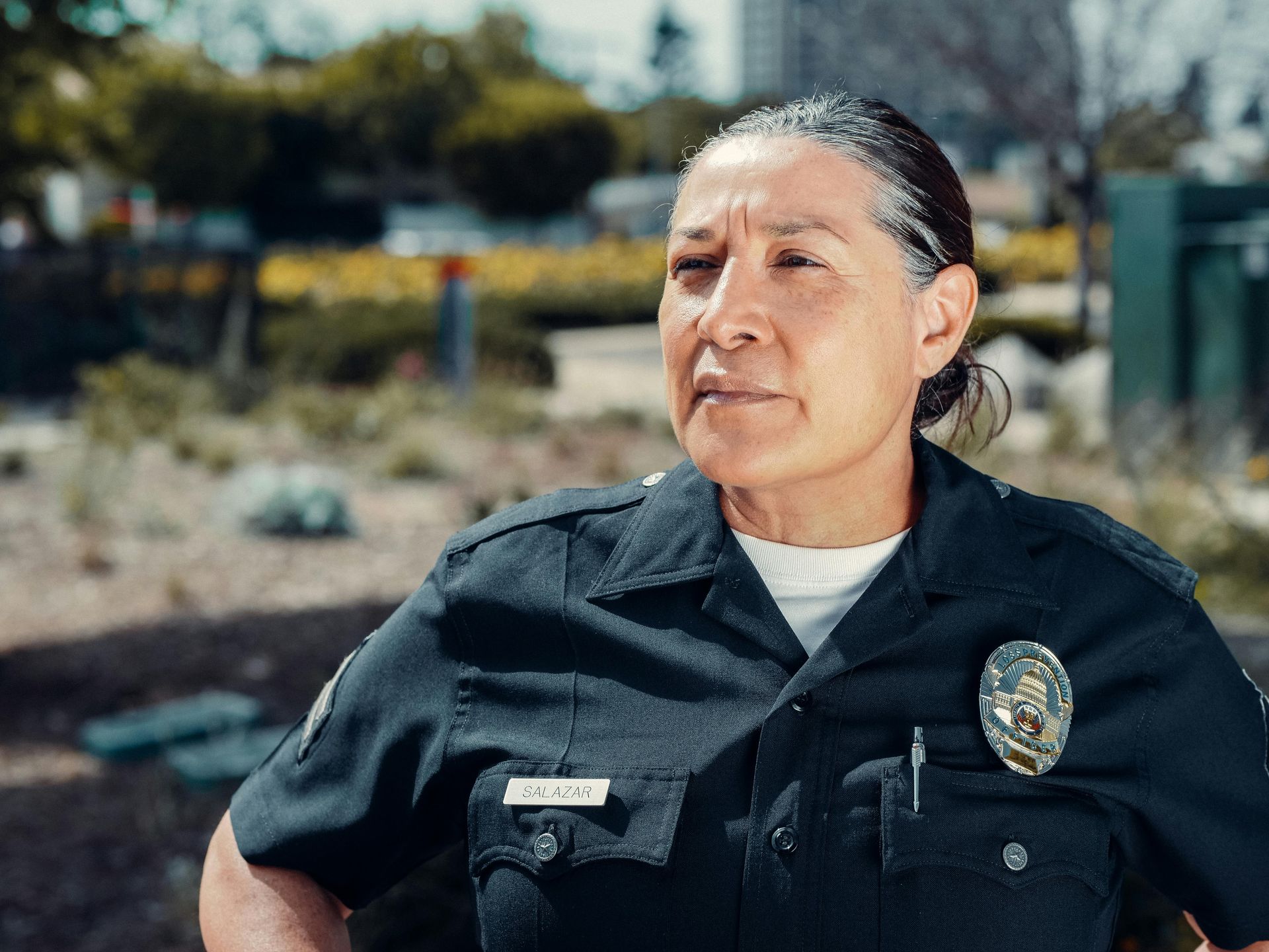 Police officer in uniform looks off-camera, hands on hips. Outdoor setting, trees, buildings in the background.