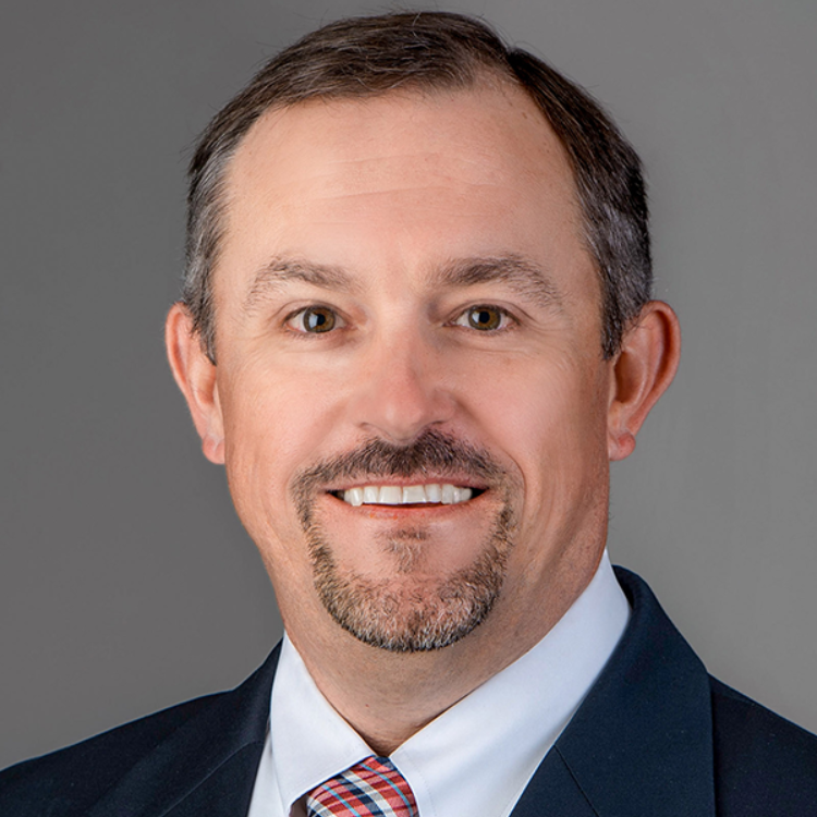 Professional headshot of a man in a dark suit and light blue shirt, smiling against a gray background