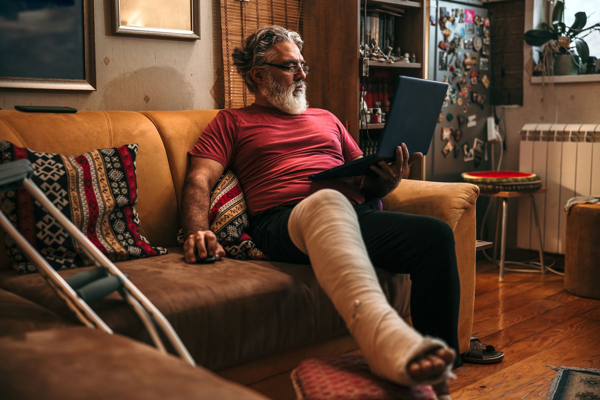Man with a broken leg sitting on a couch, using a laptop to consult with a slip and fall lawyer. Crutches are resting beside him.