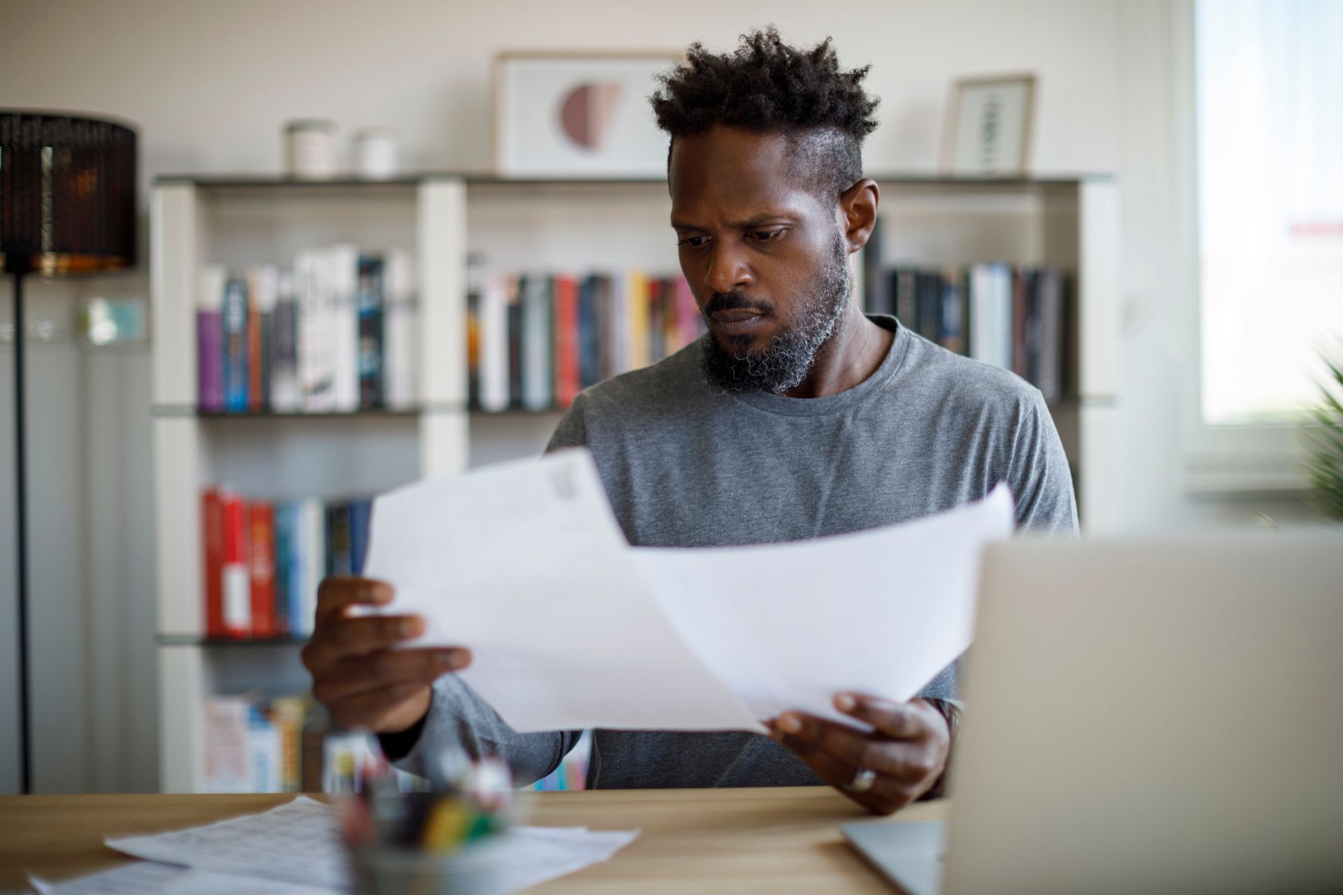A worried man sitting at a desk, carefully reading a disability benefits denial letter. A laptop and paperwork are visible in front of him.