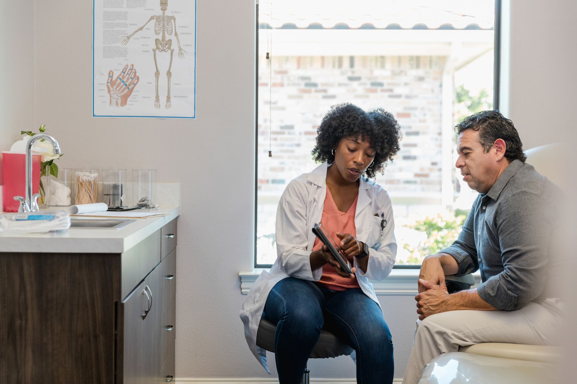 A doctor sitting in a medical office, reviewing medical records with a patient. The doctor is holding a tablet and explaining details to the patient.