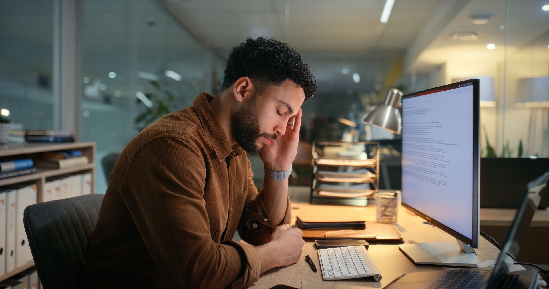 A fatigued office worker sits at a desk late at night, rubbing his forehead while looking at his computer screen, illustrating the impact of unpaid overtime.