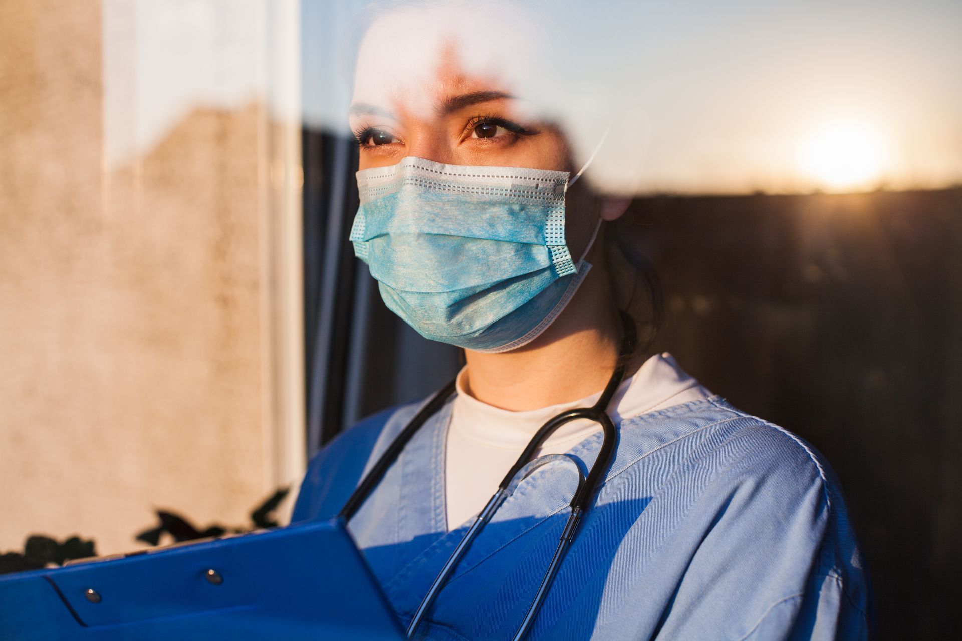 Young sad female caucasian UK US GP EMS doctor carer looking through ICU window,fear uncertainty in eyes,wearing face mask gazing at sun,hope faith in overcoming Coronavirus COVID-19 pandemic crisis.