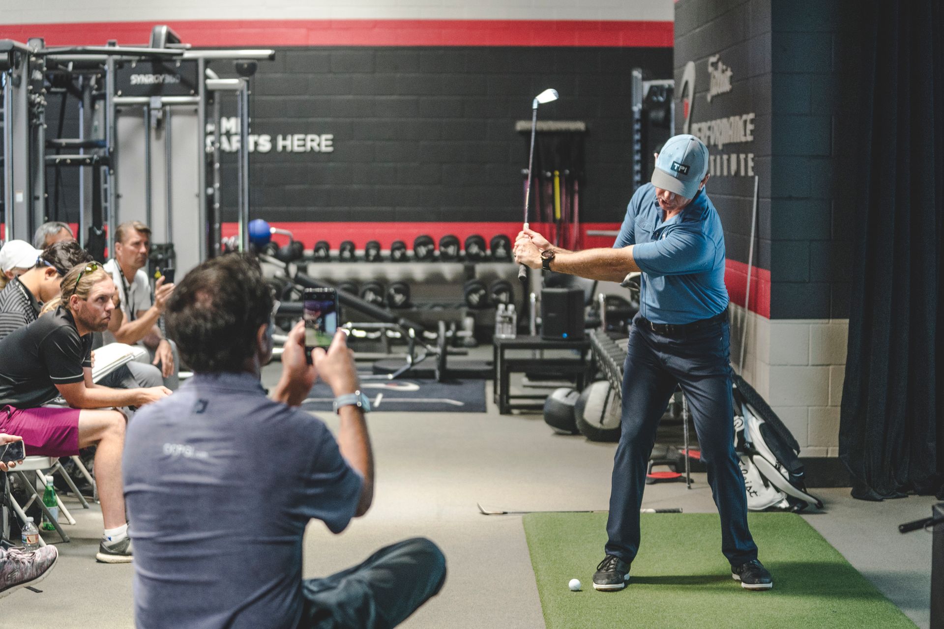 Person demonstrating a golf swing in a gym while others watch and record.