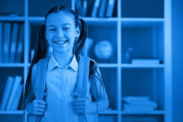 Smiling student with backpack straps stands in front of blue-lit classroom shelves