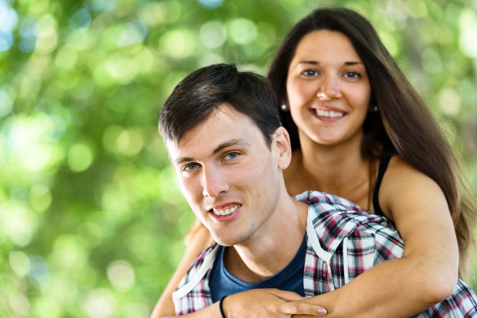Smiling couple outdoors, woman hugging man from behind against a blurred green background
