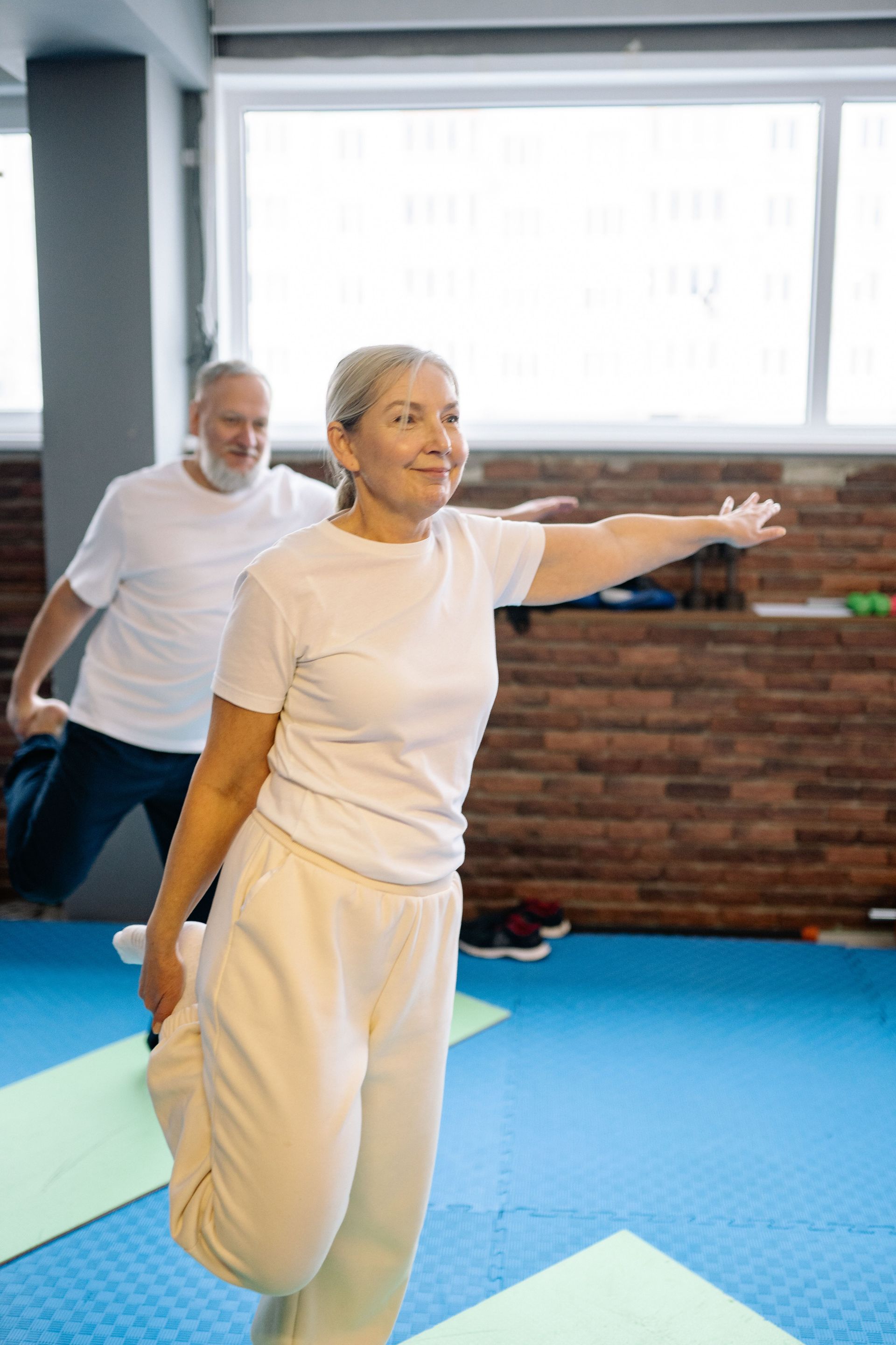 Two people stretching in a fitness studio with blue mats and a brick wall.