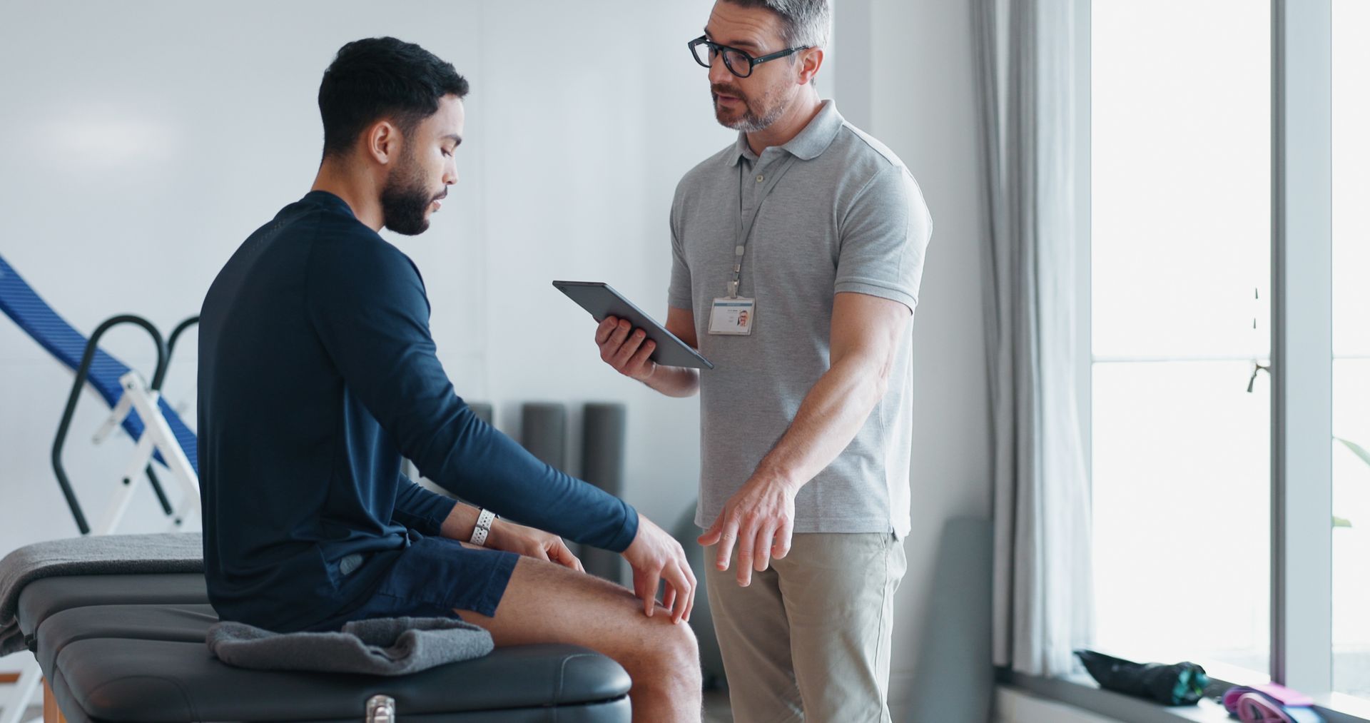 Two men talking in a clinic, one seated on a table while the other holds a tablet.
