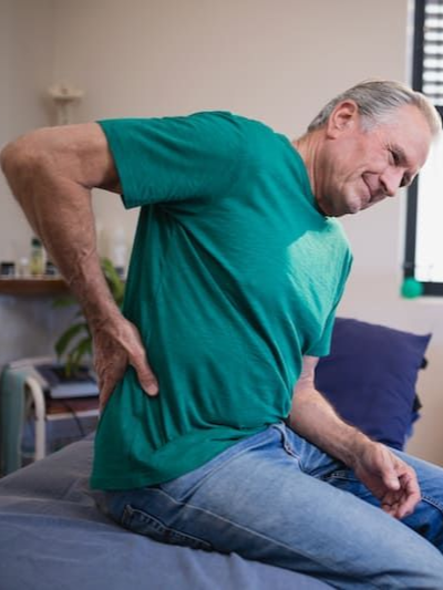 Man in a teal shirt holding his lower back while sitting on a bed, appearing to have back pain