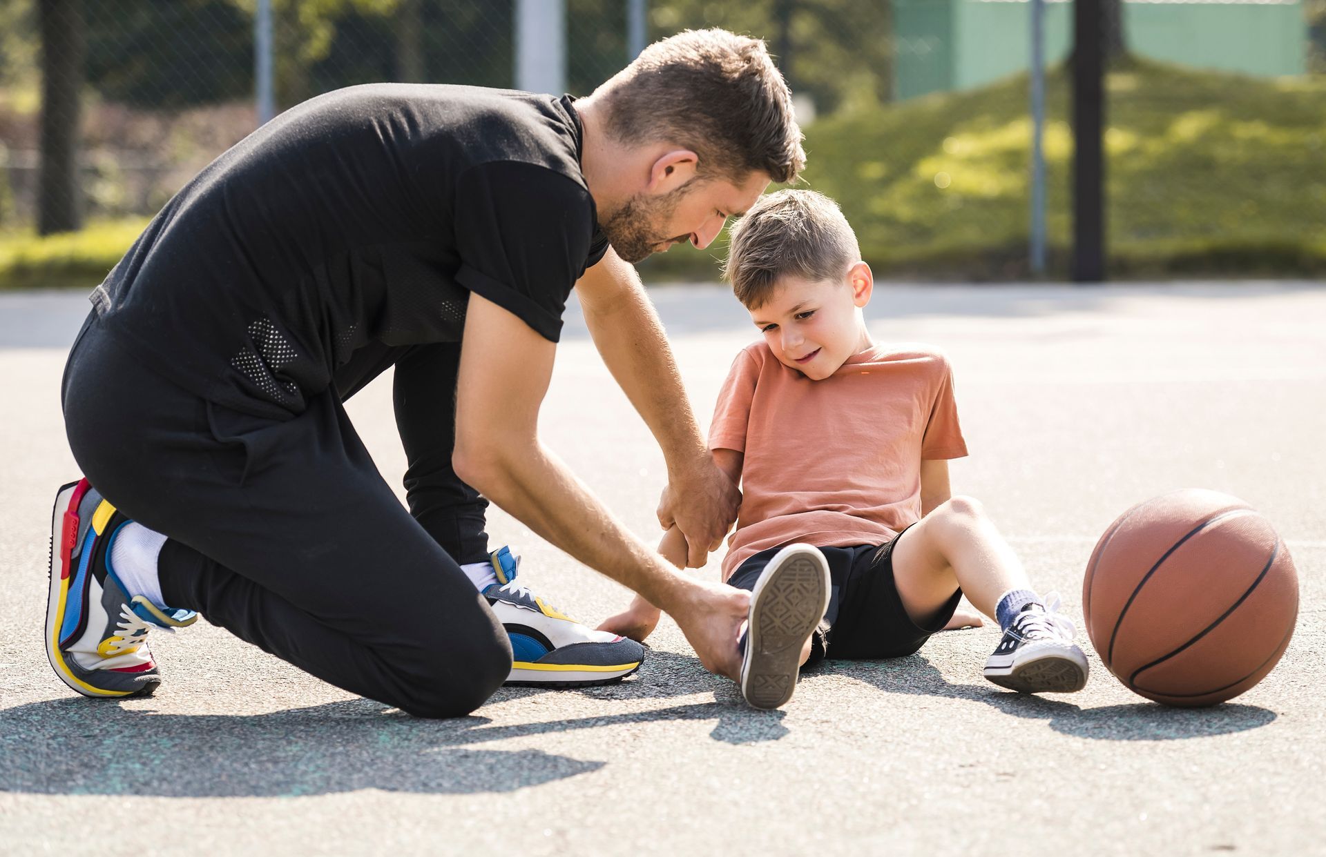 Man kneels beside a seated child on pavement, helping with a basketball outdoors