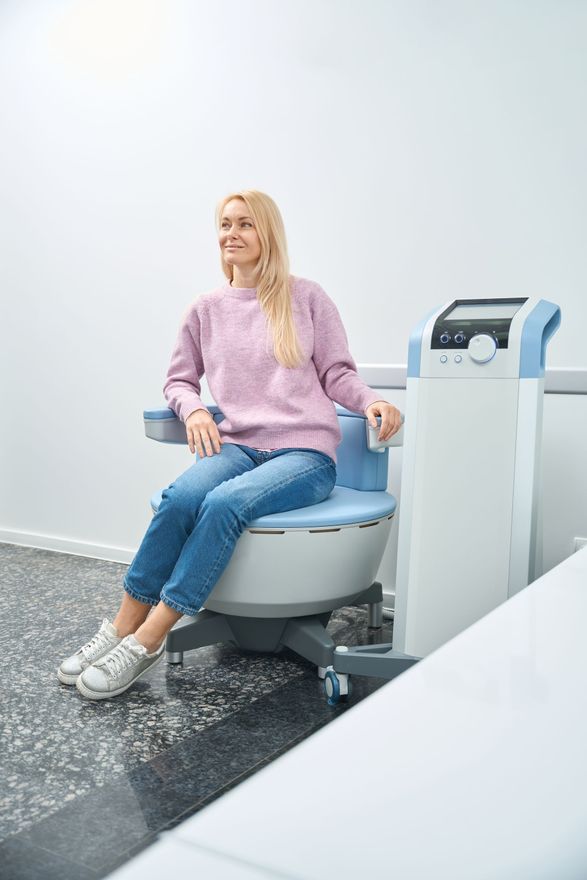 Woman seated in a clinic treatment chair beside a medical device, wearing a pink sweater and jeans.