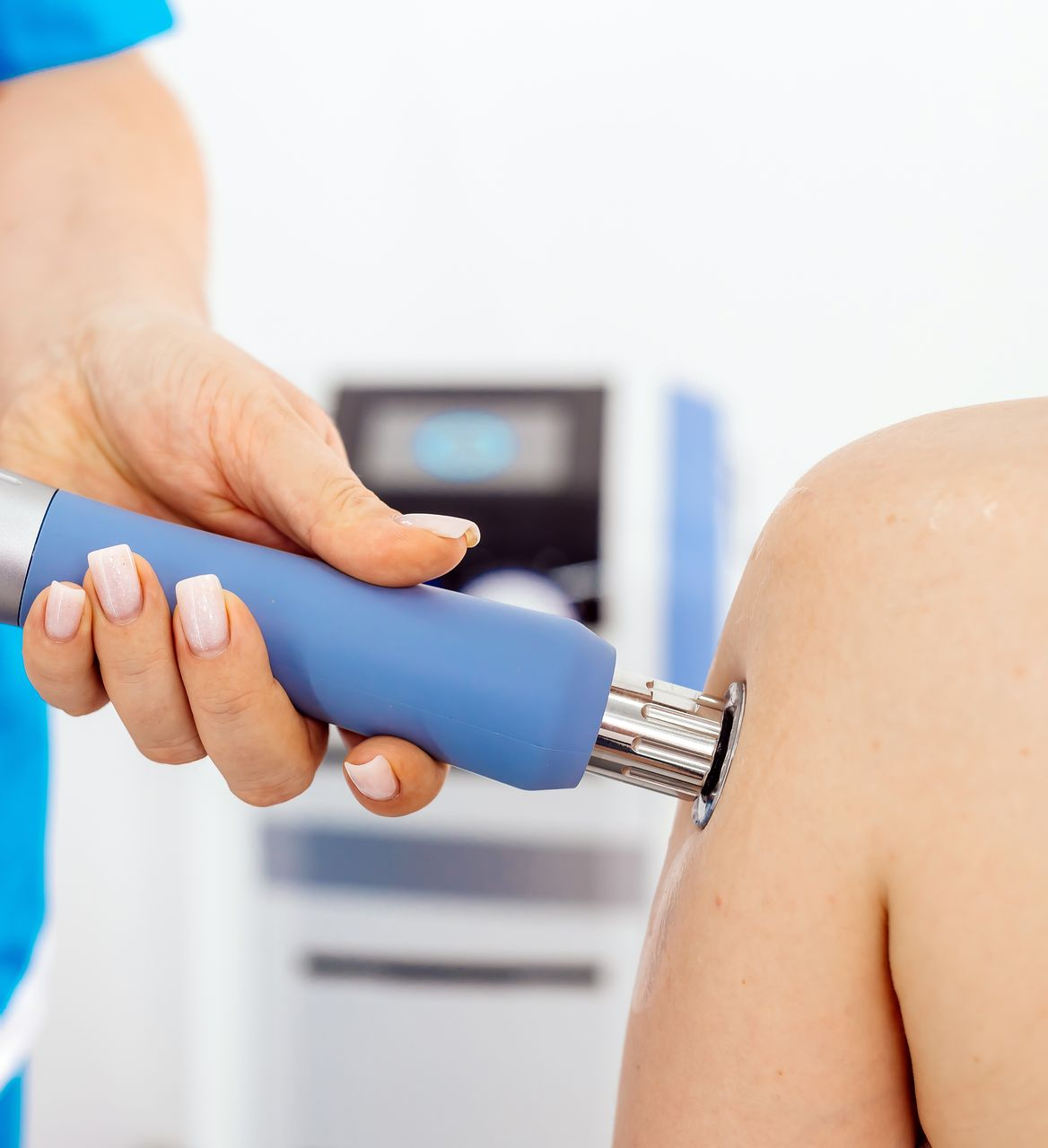 A hand holding a blue handheld device pressed against a shoulder in a medical setting